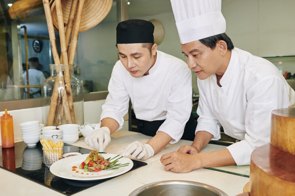 Asian mature chef teaching his student to serve the dish on the table at the restaurant