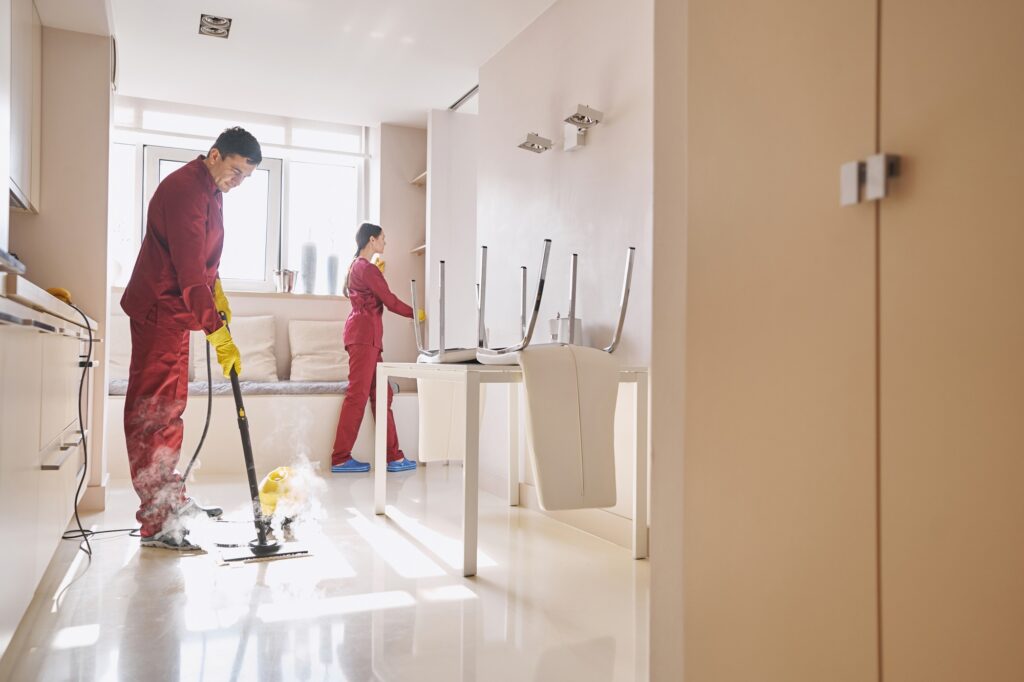 Concentrated cleaner disinfecting floor with steam mop while his coworker dusting furniture