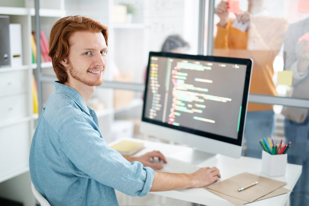 Young cheerful and successful programmer or webdesigner sitting by desk in front of computer screen and analyzing coded data